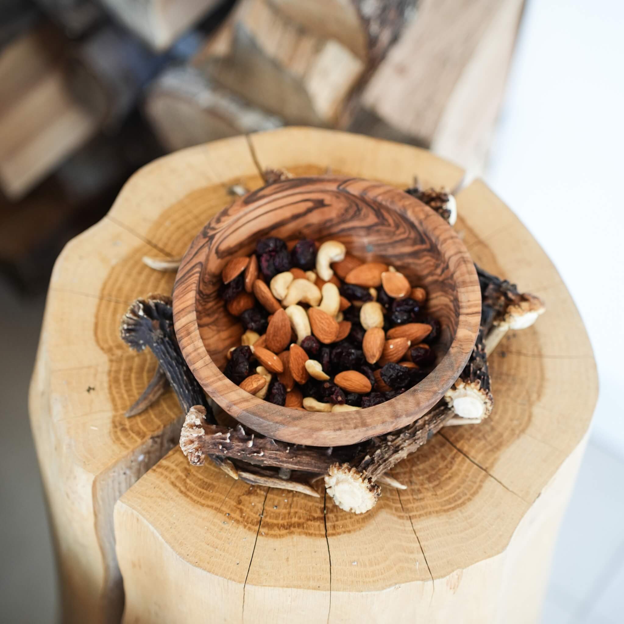 Olive wood bowl filled with mixed nuts and dried fruits, placed on a rustic wooden stump with stacked firewood in the background.