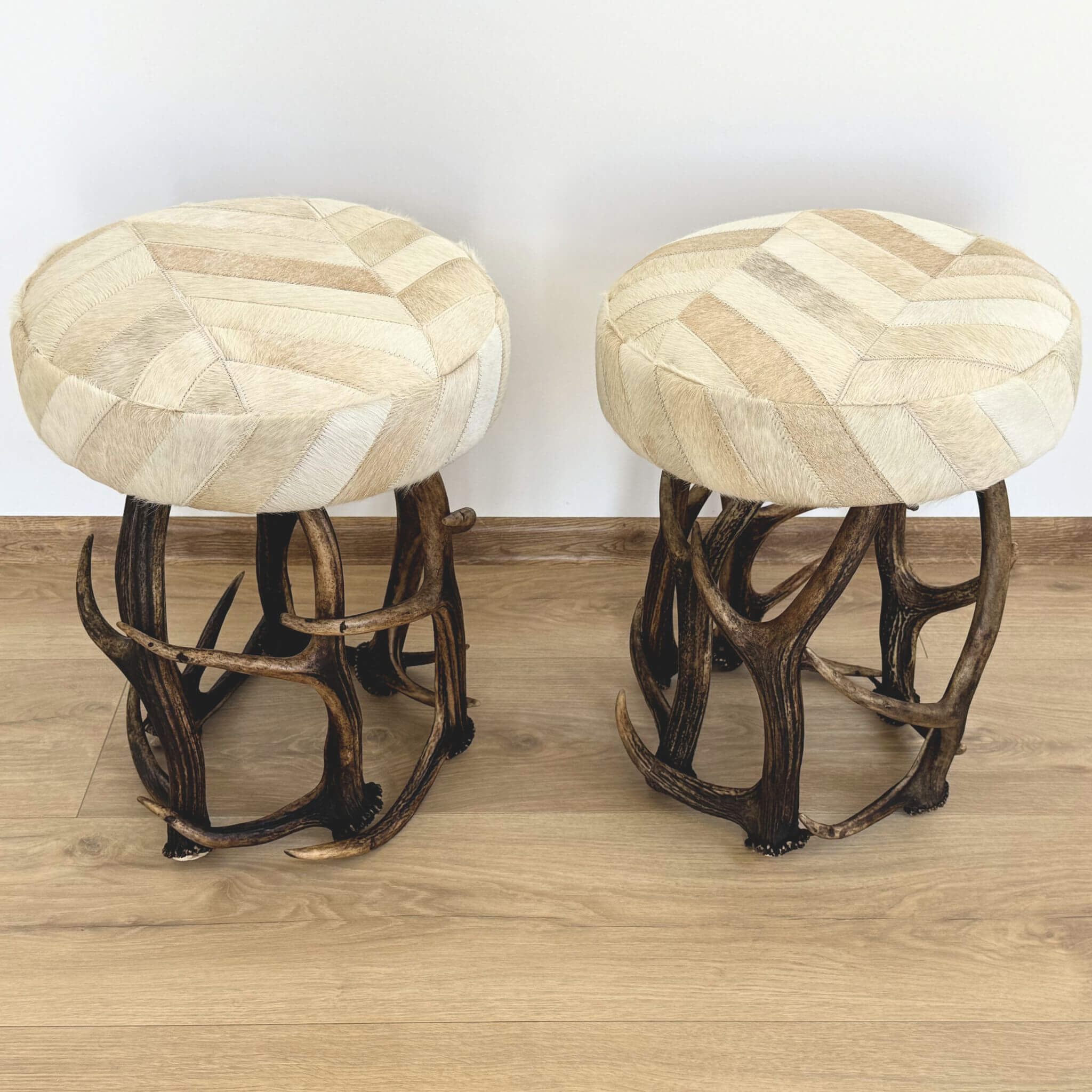 Two round upholstered stools with patchwork cowhide seats and antler-style wooden legs, shown on a light wood floor against a plain white wall.