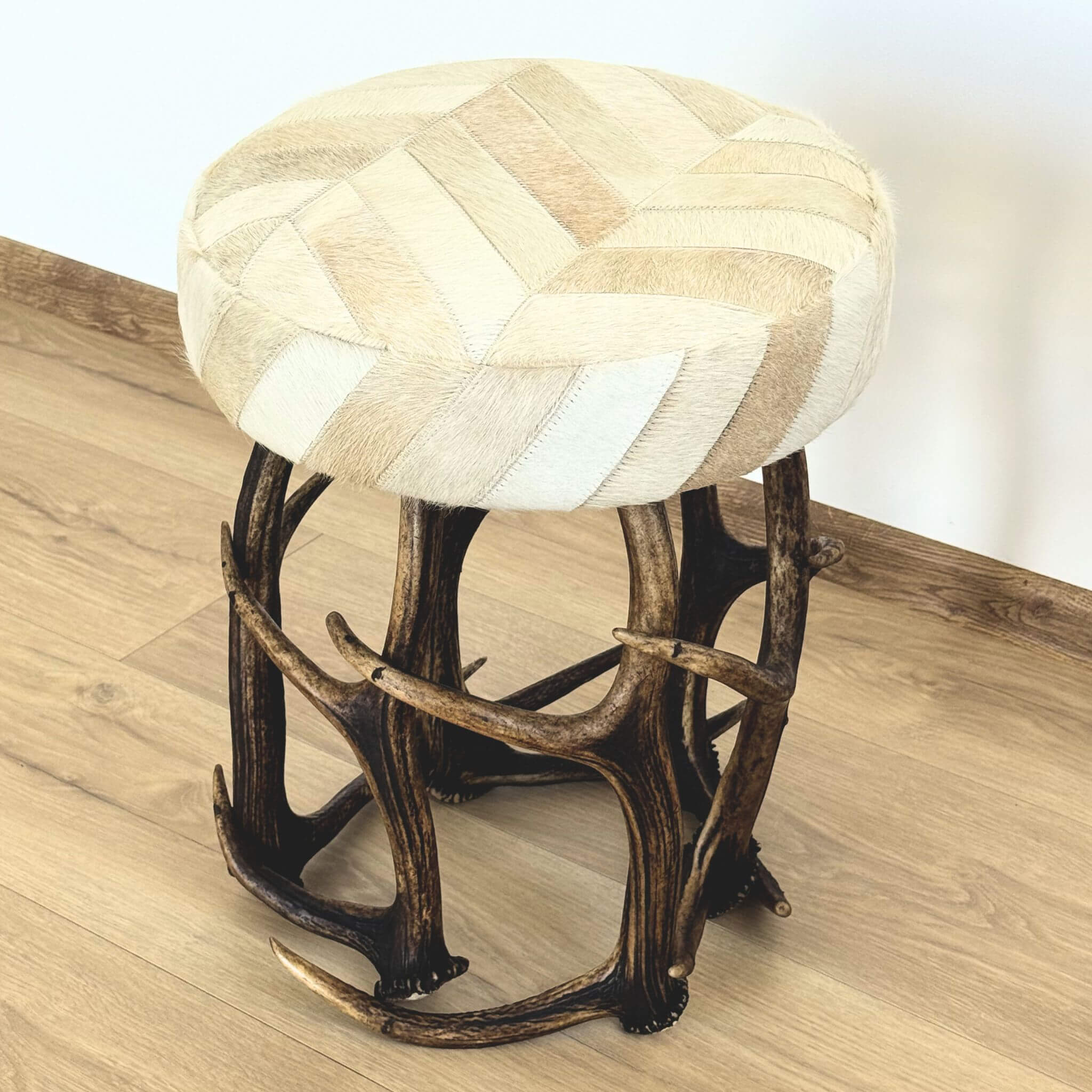 A round upholstered stool with a chevron pattern of beige, tan, and cream animal hide patches on the seat, supported by a dark brown faux-antler metal frame, set on a light wood floor.