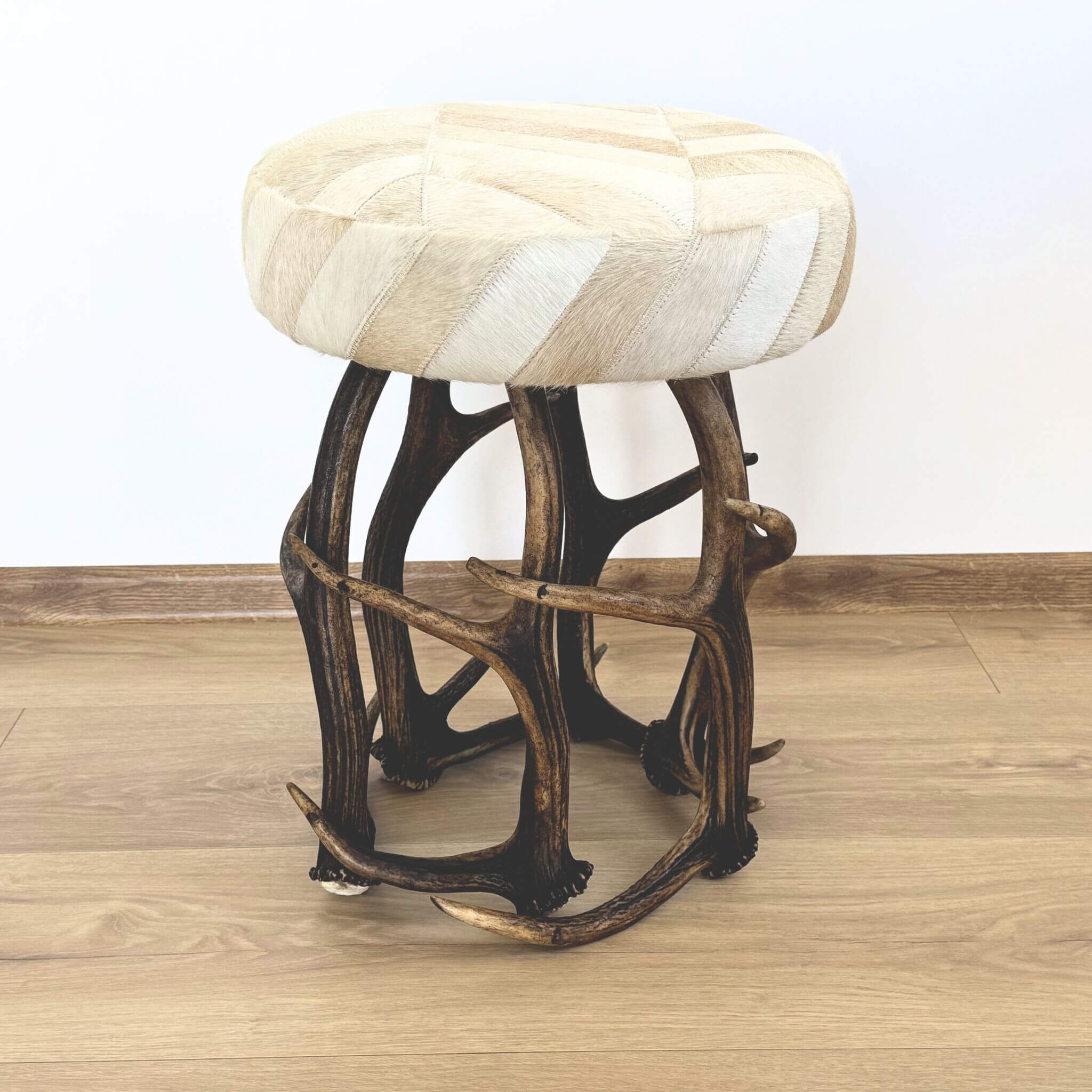 A rustic stool with a round upholstered seat in cream and beige patchwork and a dark brown antler-style metal frame, shown on a wood floor against a white wall.