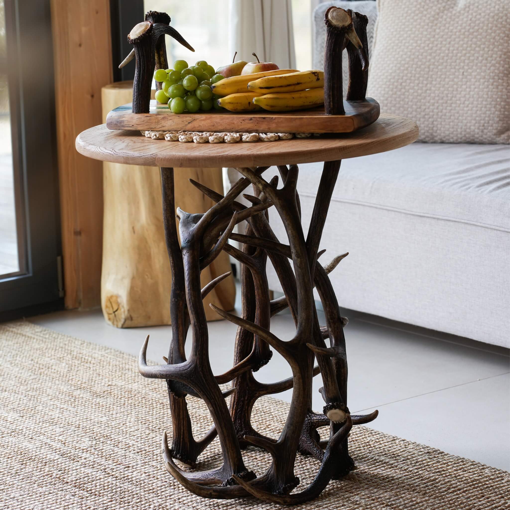 A rustic round wooden side table with a sculptural antler and branch base, styled in a living room setting with fruit on top.