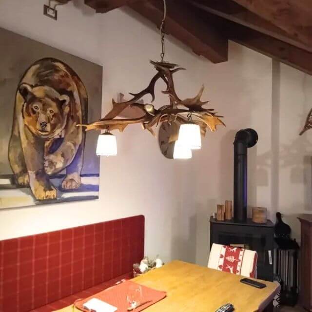 Antler pendant lamp made of fallow deer antlers. Chandelier hangs oer table in dining room. On the wall there is picture of running bear. There is fireplace in the background.