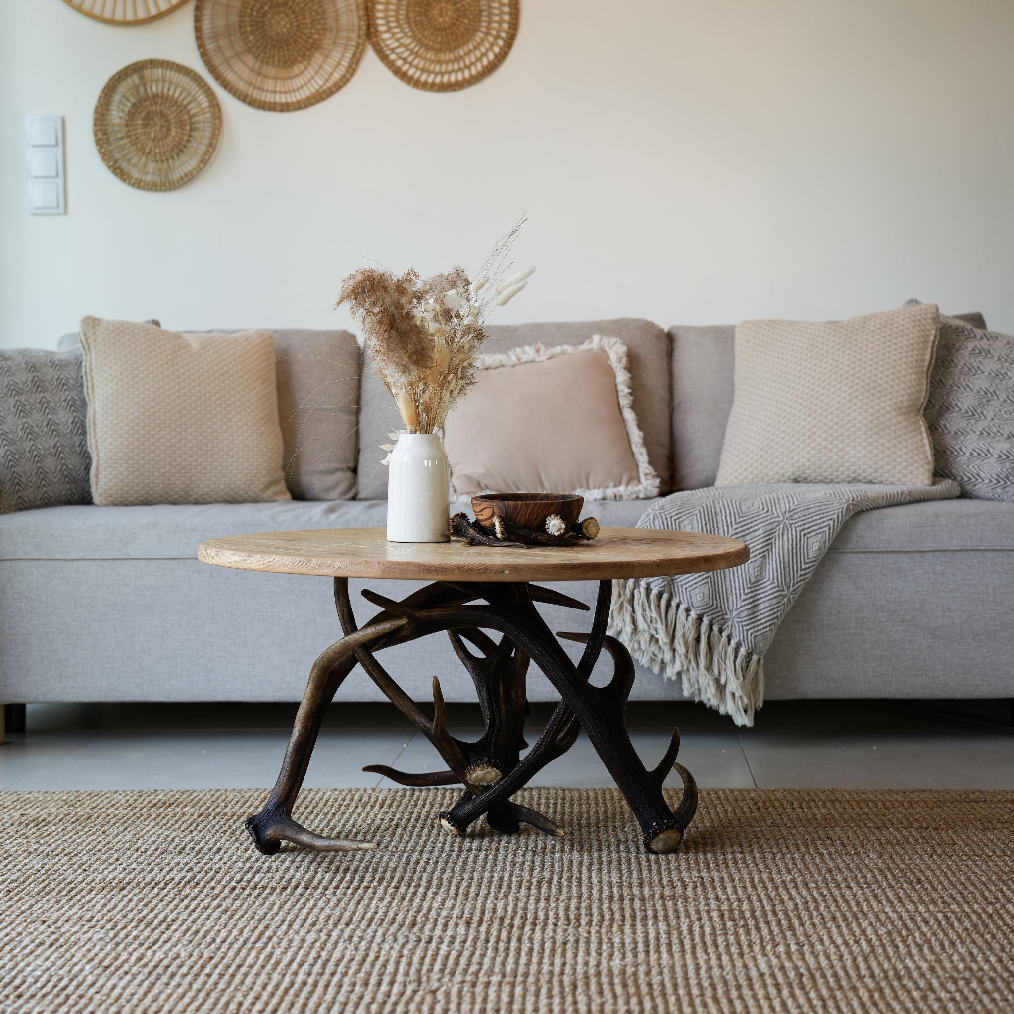 A high-angle view of a rustic, round wooden coffee table with a base made of interlocking dark deer antlers. On the table sits a white vase filled with dried pampas grass and a small wooden bowl resting on an antler piece. The table sits on a woven tan rug in front of a light gray sofa.
