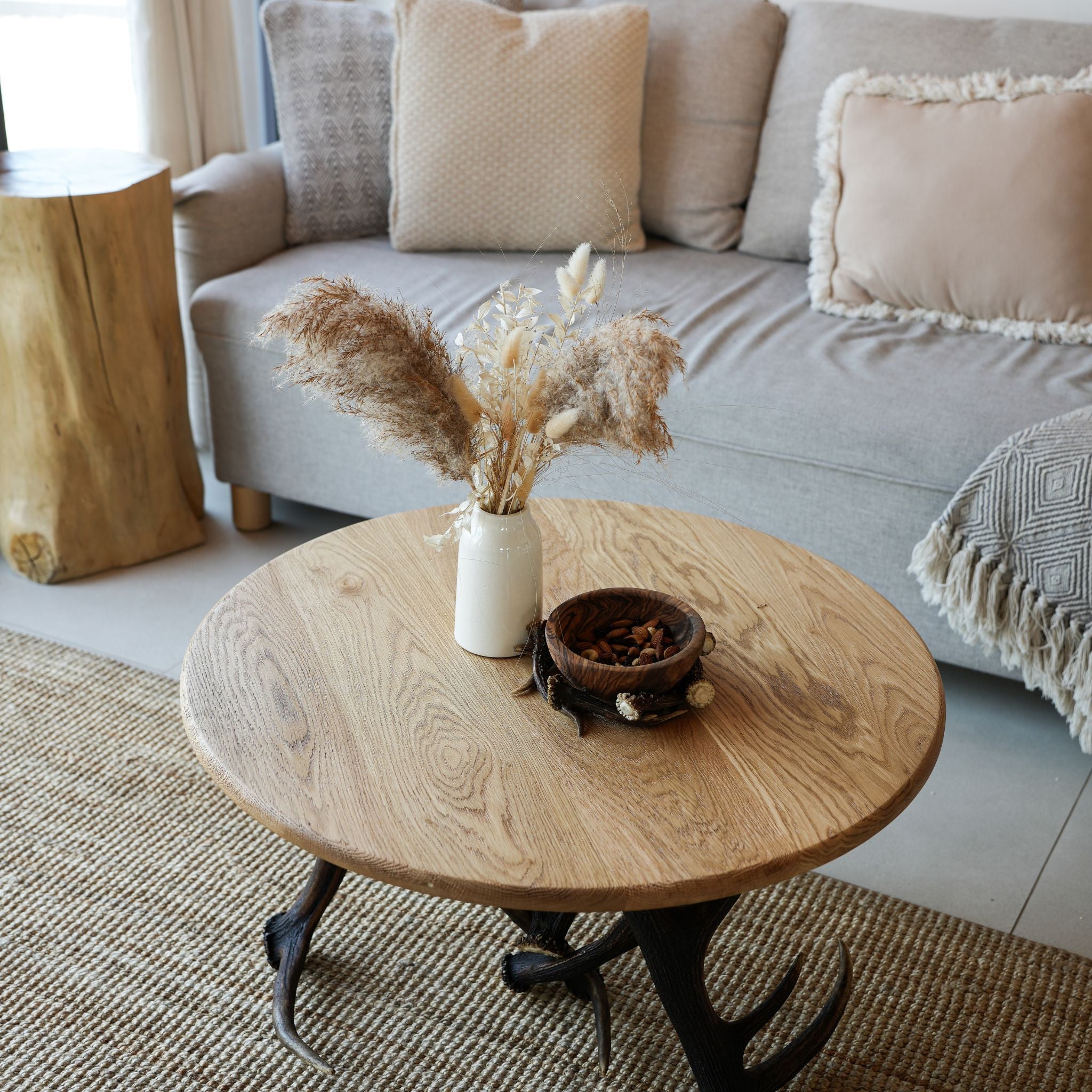 A round light oak coffee table with a dark antler-style metal base, styled in a living room with a vase of dried grasses and a small bowl on top.