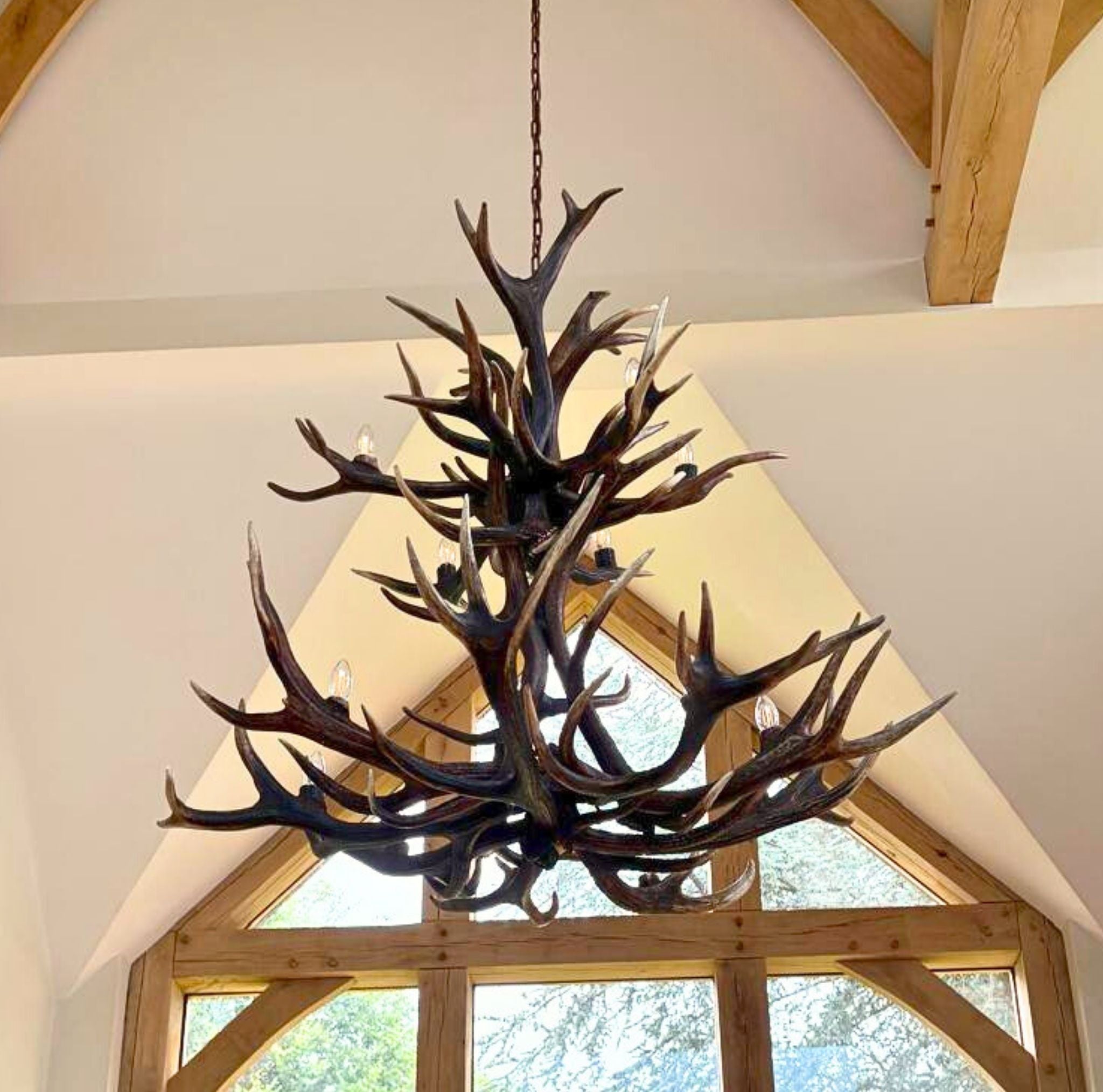 Antler chandelier hanging in a rustic room with wooden beams and large windows.