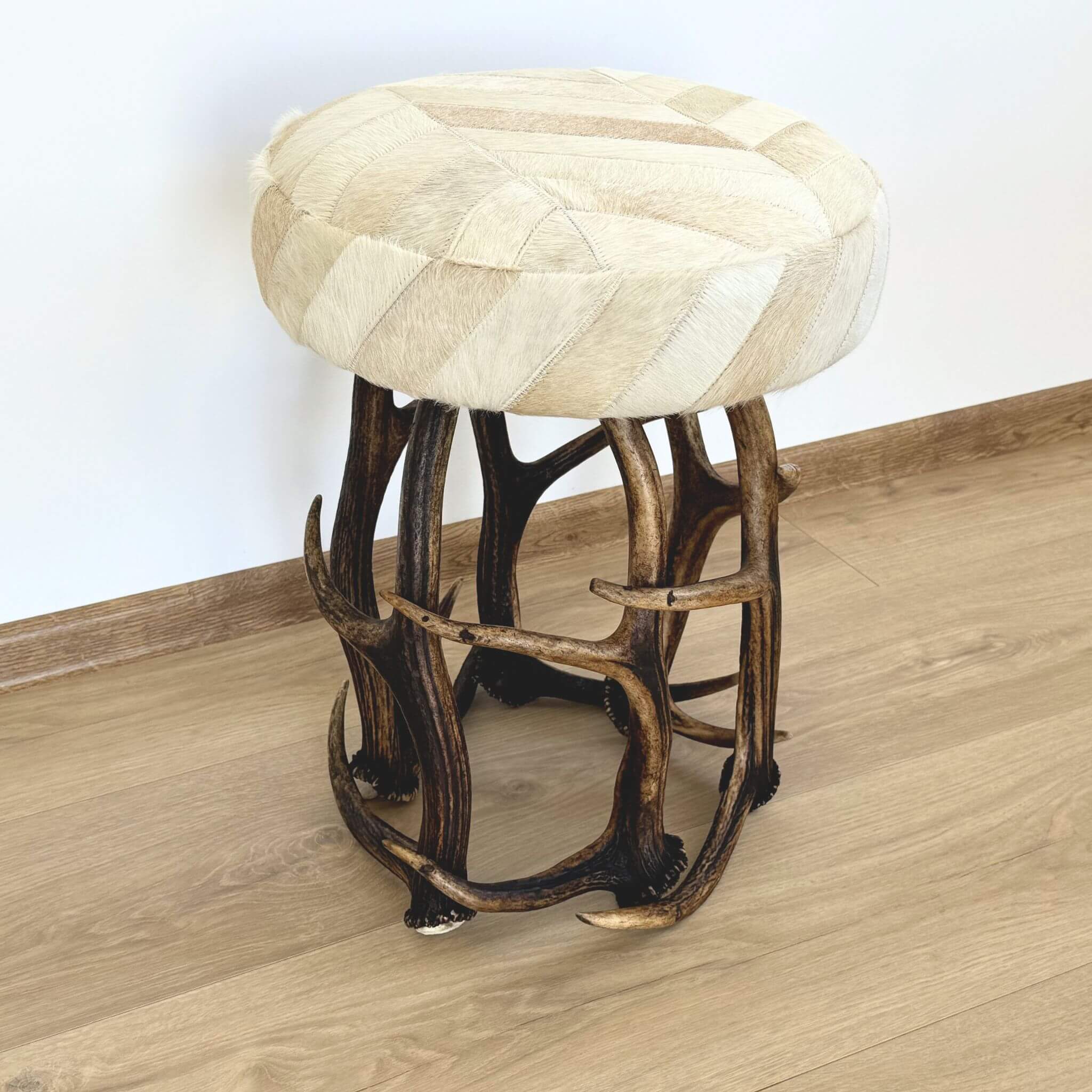 A round upholstered stool with a cream and beige patchwork hide seat and a dark brown antler-style metal base, shown on a wood floor against a white wall.