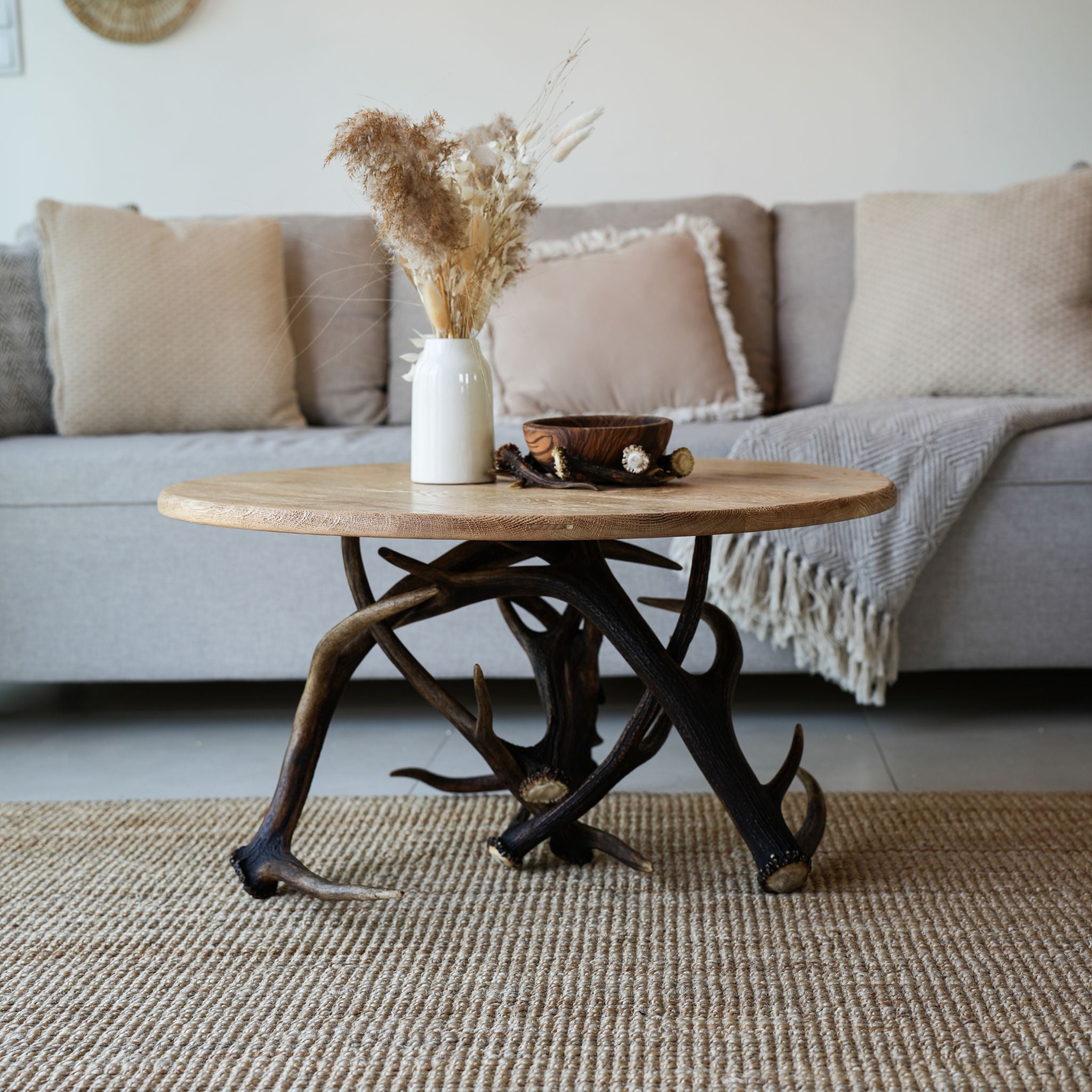 A round wooden coffee table with a dark antler-style base, styled in a living room setting with a gray sofa, beige throw pillows, a white vase with dried grass, and a small wooden bowl on top.