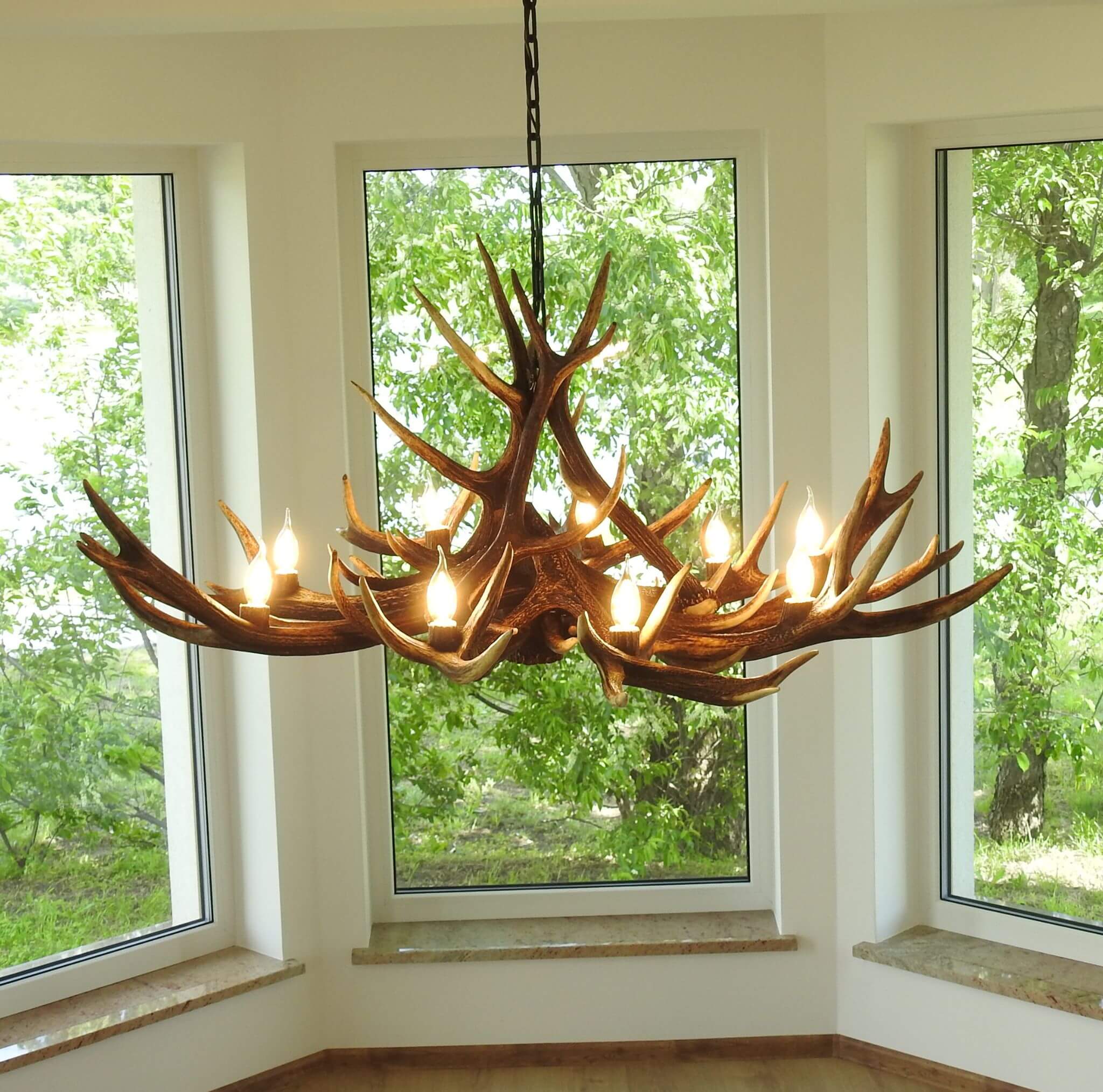 Antler chandelier hanging in a room with large windows showing greenery outside.