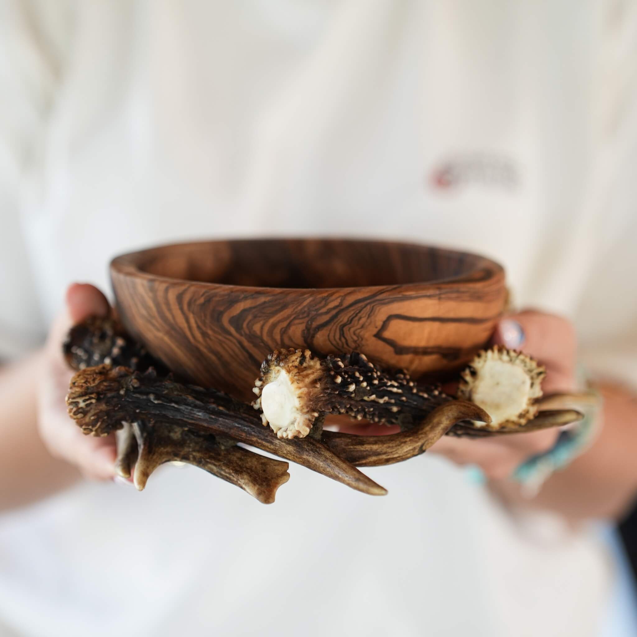 Person presenting a decorative olive wood bowl surrounded by antlers, containing dried fruit and nuts.