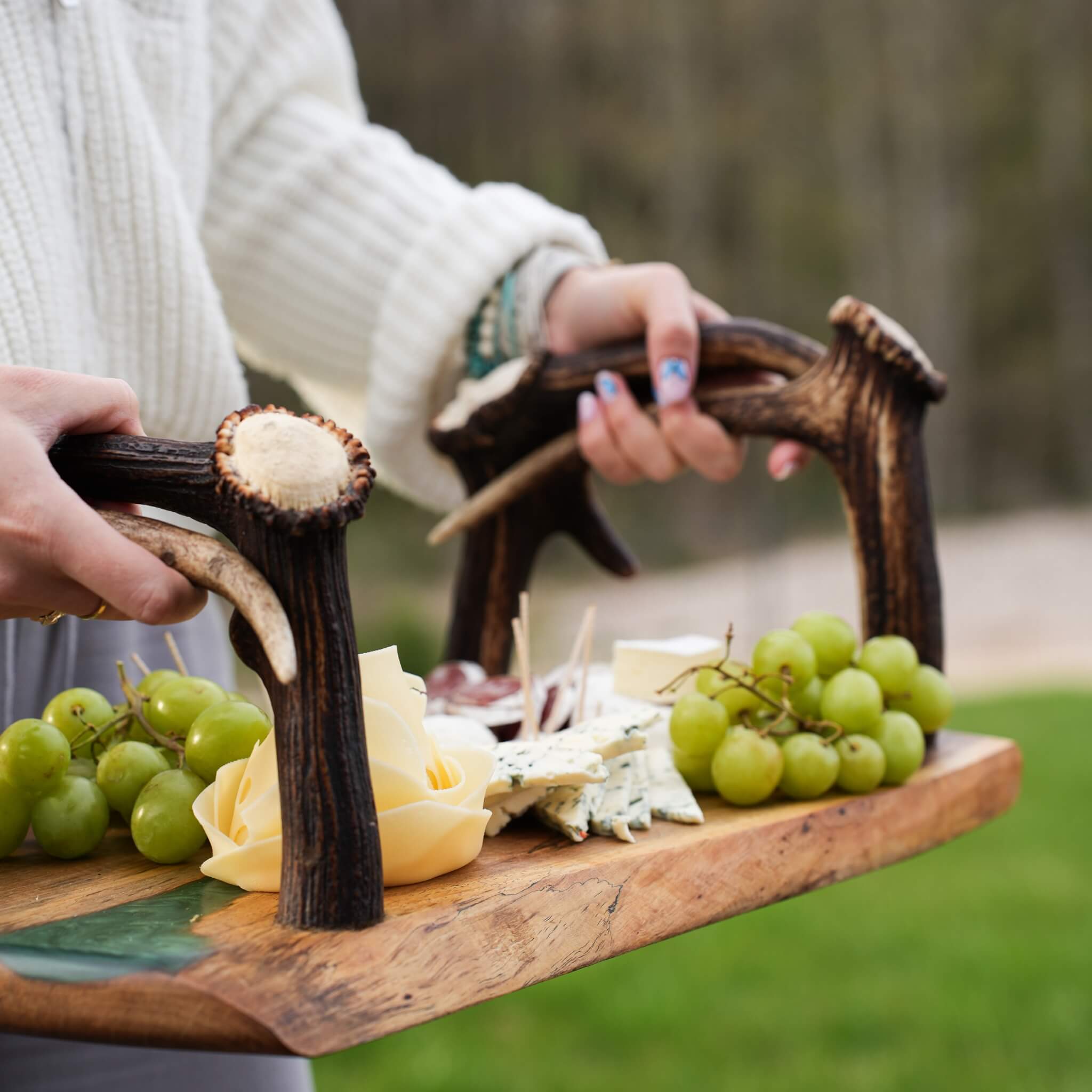 Person holding rustic olive wood tray with epoxy and antler handles, serving cheese and grapes outdoors.