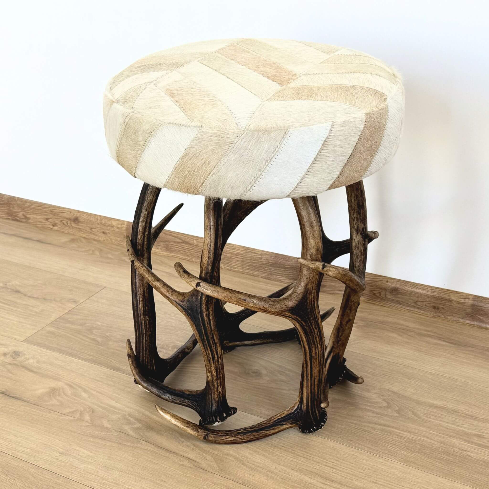 A round rustic stool resting on a light wooden floor against a white wall, featuring a base made of intricately interwoven brown antlers and a seat upholstered in light-colored animal hide arranged in a herringbone pattern.