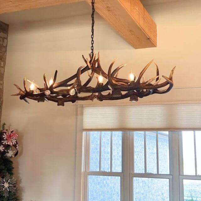 Large, elliptical shape real antler chandelier hangs in living room over the table. Chandelier hangs in rustic home on wooden beam.