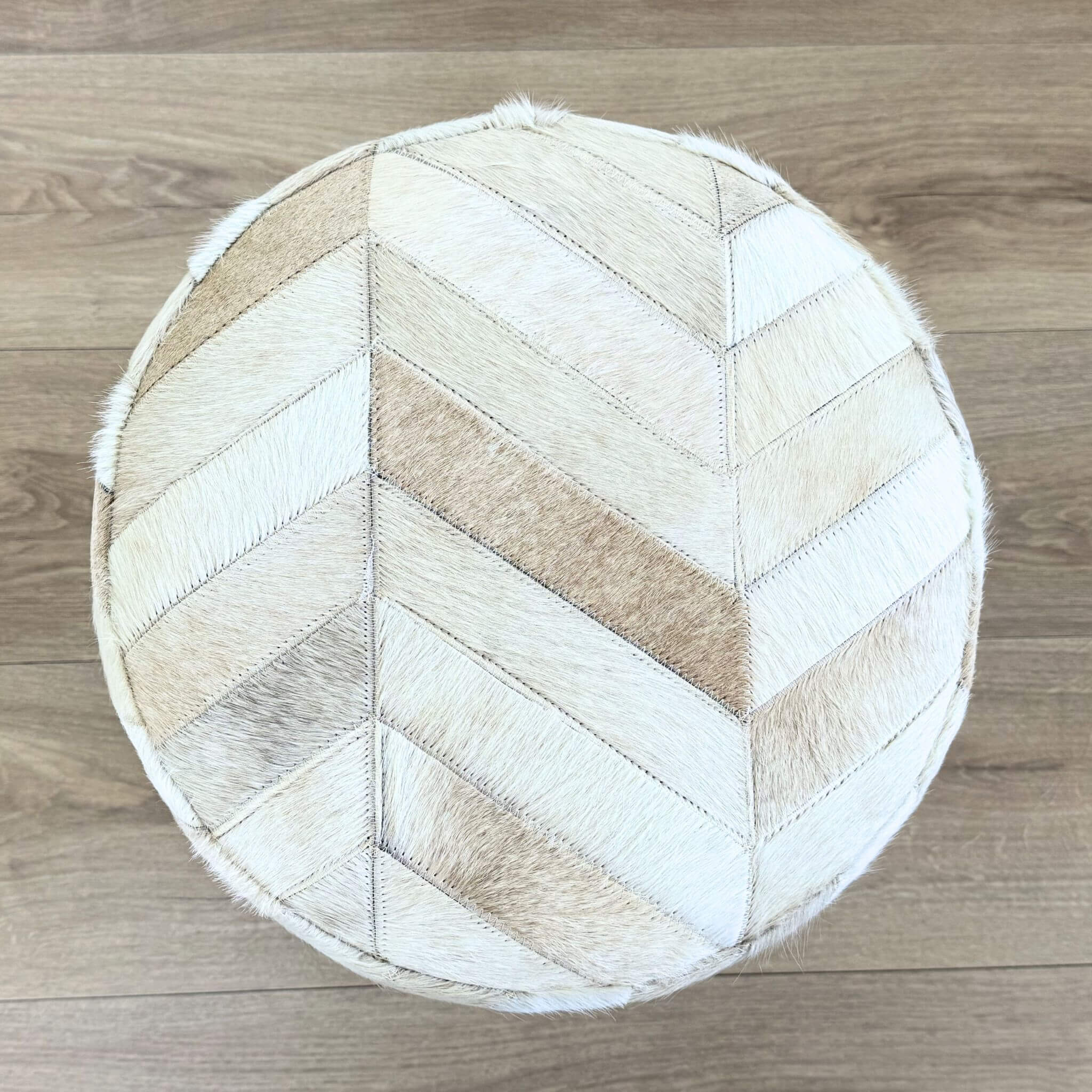 A round pouf ottoman upholstered in a chevron pattern of natural cowhide patches in cream, beige, and light brown, photographed from directly above on a light wood floor.