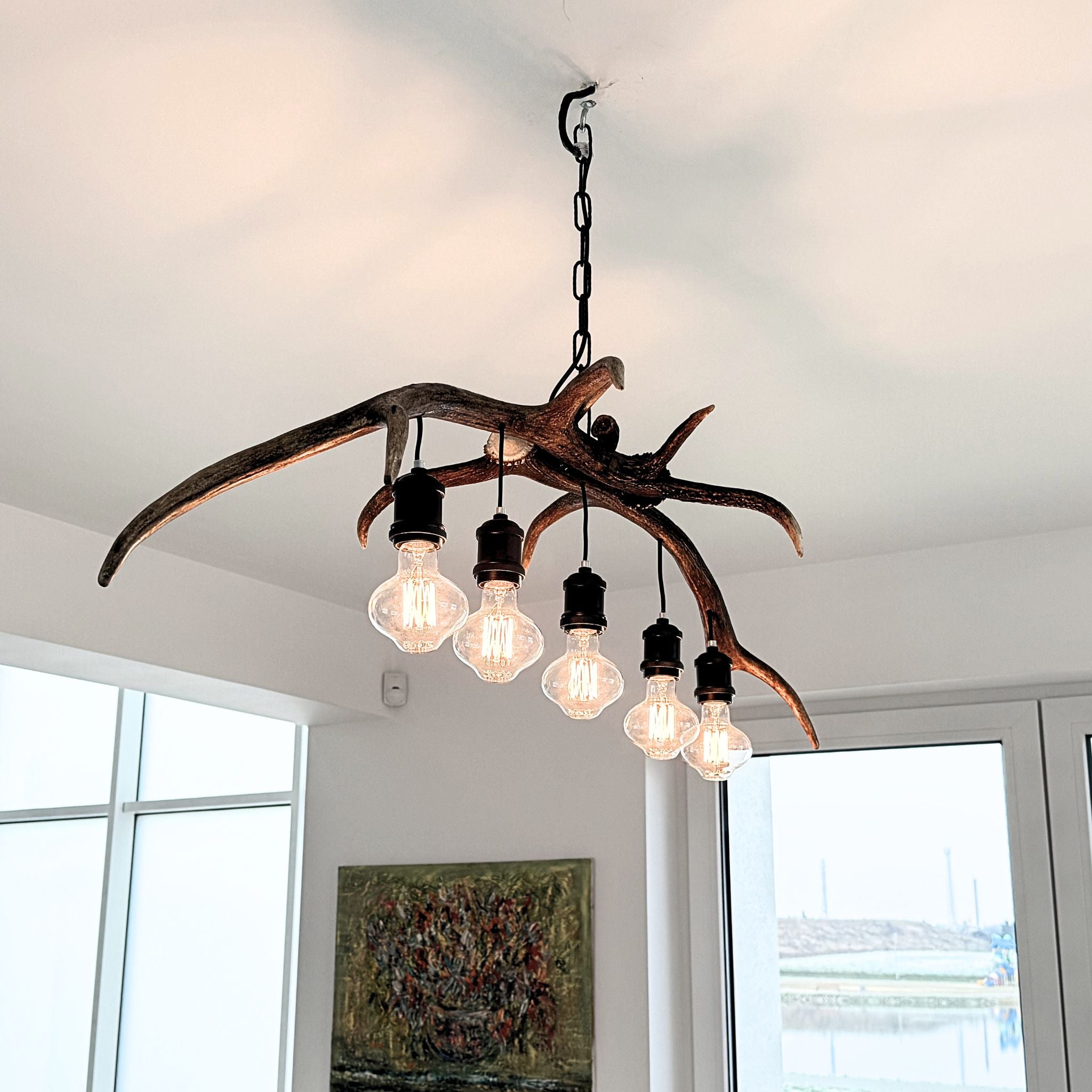 A rustic antler-style chandelier with five hanging Edison bulbs, suspended from a ceiling in a bright room with windows and a framed painting below.