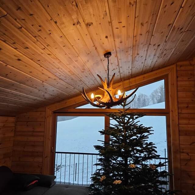 Real antler chandelier hangs in rustic home in the mountain. By the chandelier there is christmas tree.
