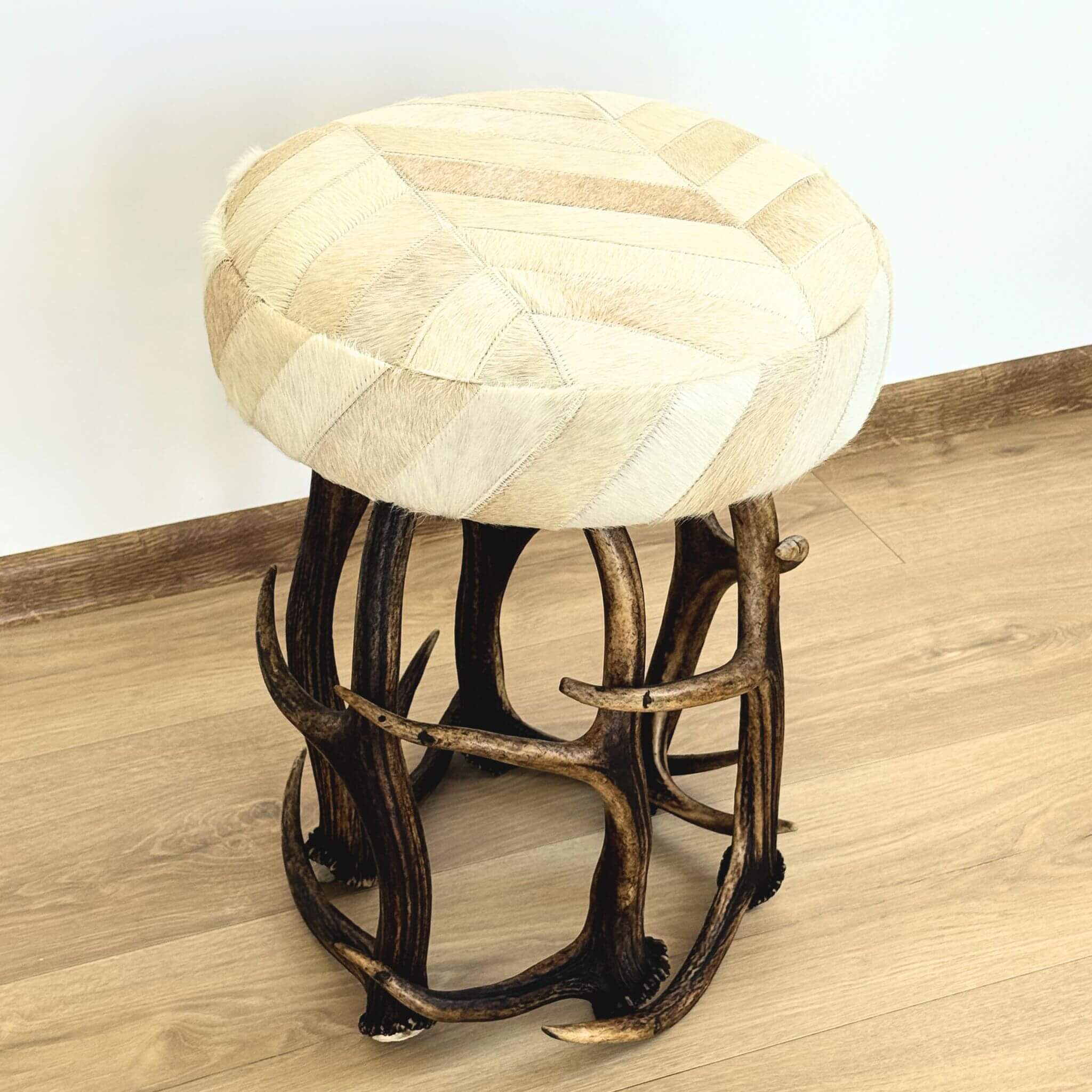 A round upholstered stool with a cream and beige patchwork cowhide seat and a dark brown antler-style metal base, shown on a wood floor against a plain wall.