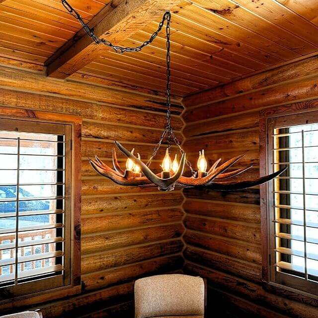 Small antler chandelier hanging on chain in a rustic wooden cabin with large windows.