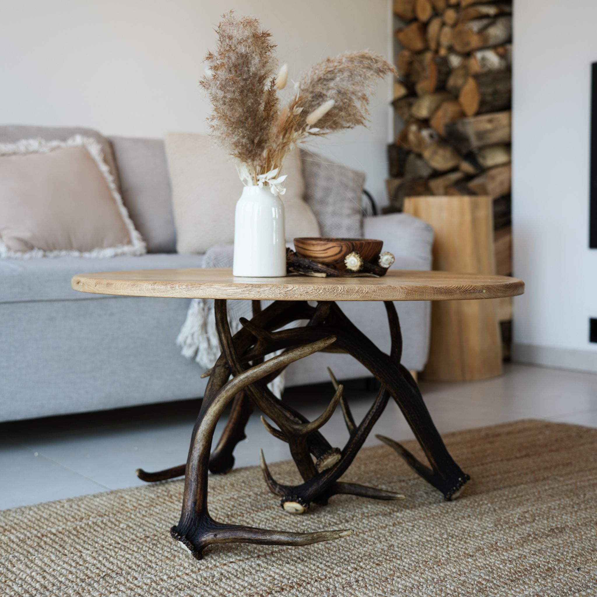 A rustic coffee table with a round light wood top and a dark natural antler-style base, styled in a living room with a white vase of pampas grass and a small bowl on top.