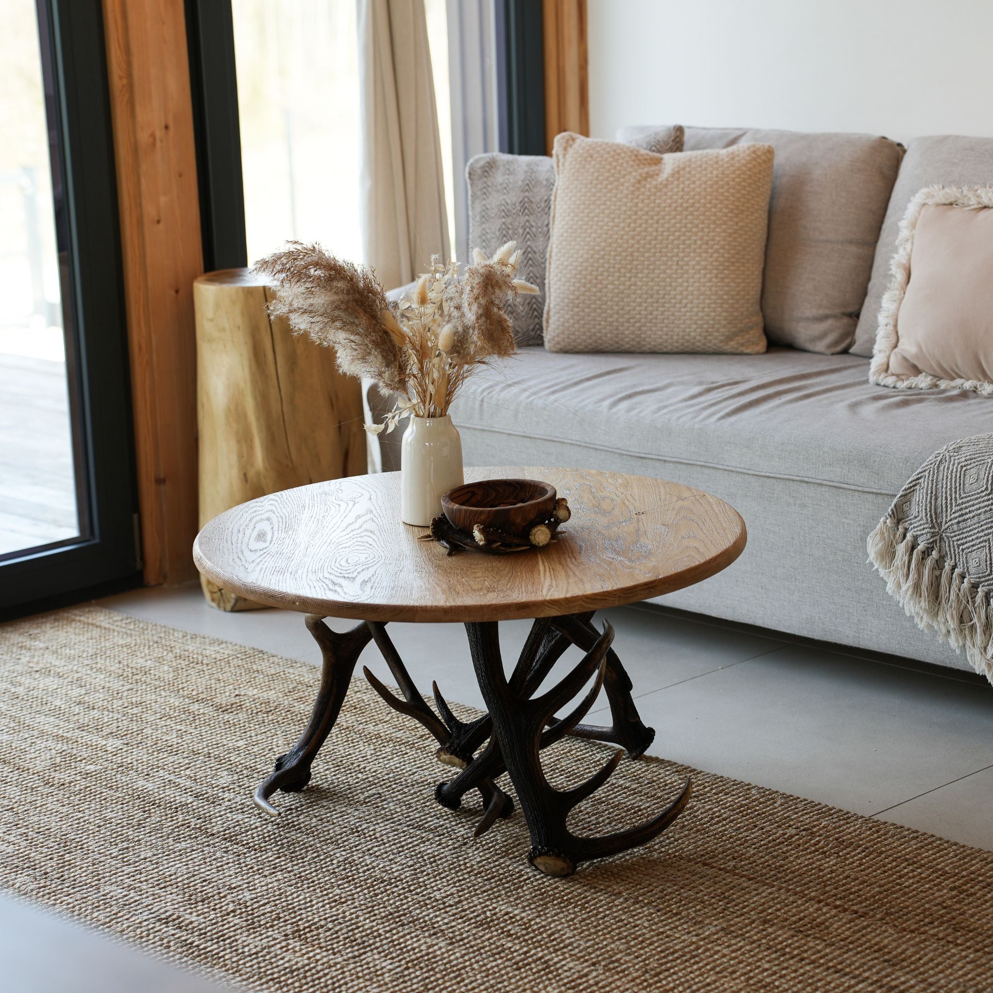 A round wooden coffee table with antler-style legs in a modern living room setting, styled with a small vase of pampas grass and a decorative bowl.